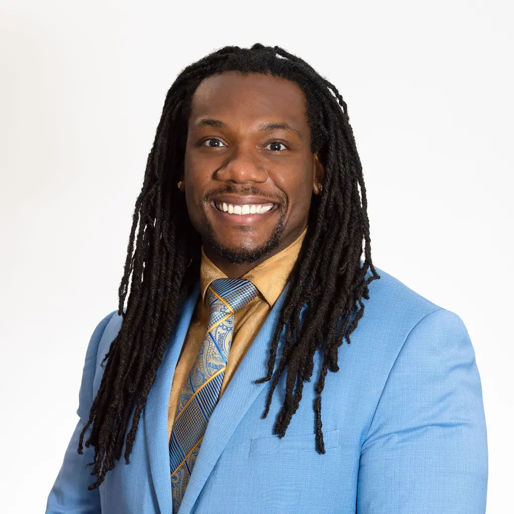 A man with dreads in a blue suit and yellow tie.